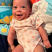 Legacy is registered to the contest to win money with this photo: baby, infant, smiling, happy, cute, face, child, onesie, patterned_clothing, curly_hair, skin, lying_down, blanket, colorful, soft_toys, comfort, portrait, indoors, closeup, expression