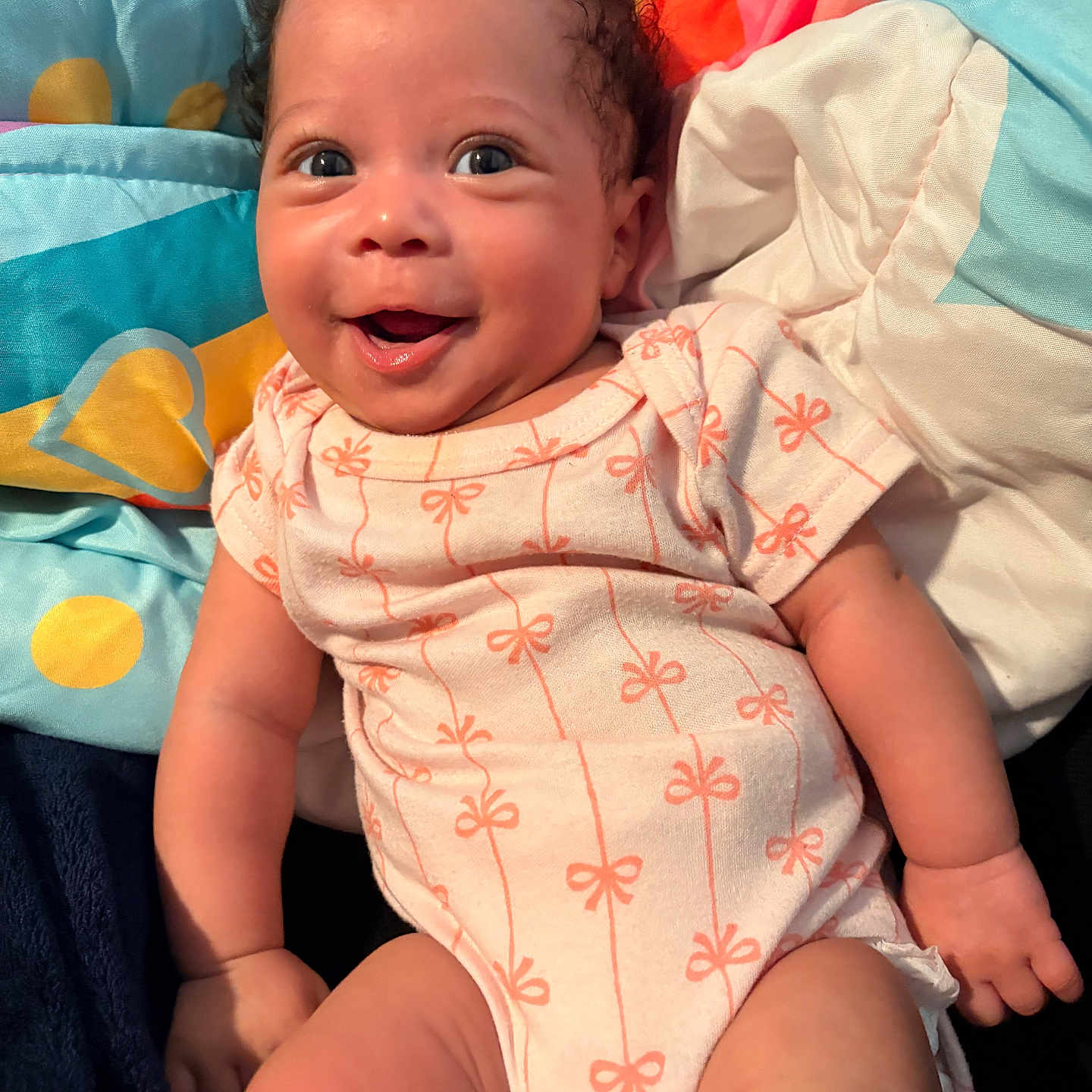 Legacy is registered to the contest to win money with this photo: baby, infant, smiling, happy, cute, face, child, onesie, patterned_clothing, curly_hair, skin, lying_down, blanket, colorful, soft_toys, comfort, portrait, indoors, closeup, expression