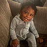 child, toddler, smiling, portrait, sitting, couch, indoors, tracksuit, gray_clothing, hoodie, curly_hair, happy, face, teeth, socks, furniture, living_room, cozy, candid, photography