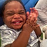 Bryson is registered to the contest to win money with this photo: baby, child, clapping, smiling, laughing, portrait, close_up, face, teeth, curly_hair, happy, joyful, indoors, couch, adult_hand, patterned_clothing, blue_background, sitting, family, photography
