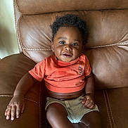Bryson joined the competition — help win amazing prizes! baby, child, infant, sitting, couch, leather, portrait, smiling, curly_hair, coral_shirt, shorts, indoors, furniture, brown, casual, front_facing, relaxed, adorable, photography, home