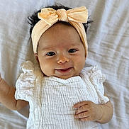 Nara is registered to the contest to win money with this photo: baby, infant, headband, bow, smile, white_clothing, textured_fabric, ruffles, lying_down, blanket, soft, cute, portrait, child, happy, face, hand, skin, indoors, person
