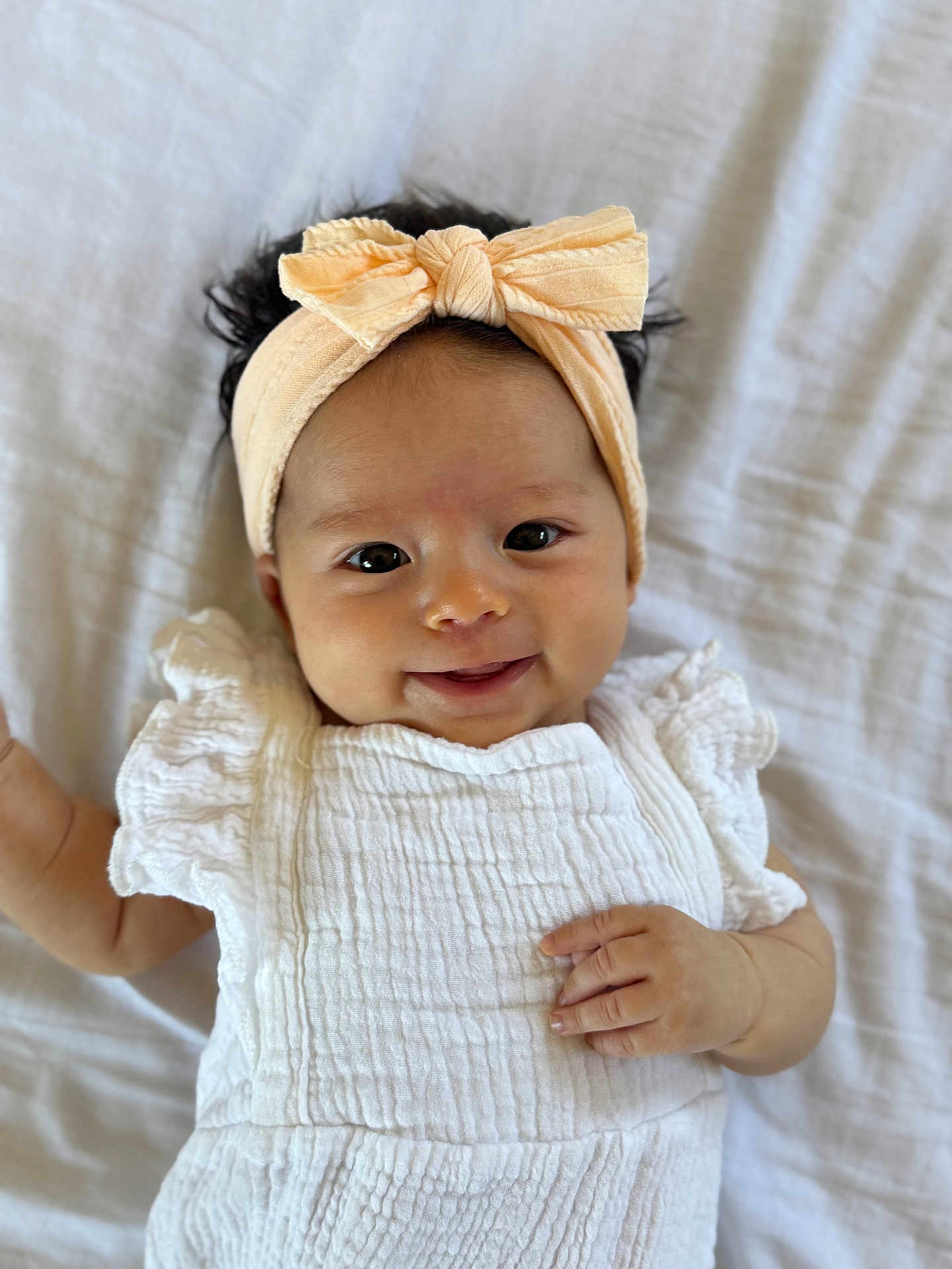 Nara is registered to the contest to win money with this photo: baby, infant, headband, bow, smile, white_clothing, textured_fabric, ruffles, lying_down, blanket, soft, cute, portrait, child, happy, face, hand, skin, indoors, person