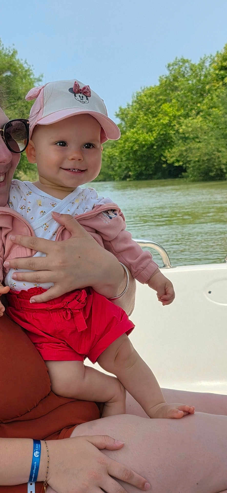 Elina a rejoint le concours — aidez-le/la à gagner de superbes lots ! baby, child, smiling, cap, pink, white, red_shorts, outdoor, river, trees, greenery, adult, holding, boat, water, nature, sunlight, happy, person, summer