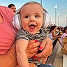 baby, headphones, striped_clothing, smiling, happy, person, adult_hand, nail_polish, outdoor, bench, spectators, evening, light_poles, crowd, casual_wear, closeup, infant, joy, parent, fun