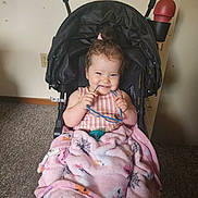 Alice is registered to the contest to win money with this photo: toddler, child, stroller, blanket, glasses, indoor, carpet, wall, filing_cabinet, pink_blanket, smiling, happy, baby, clothing, toy, cozy, sitting, person, cute, playful