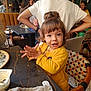 toddler, child, yellow_jacket, clapping, table, cafe, chair, people, indoor, happy, smiling, curly_hair, person, food, plate, spoon, warm_lighting, casual, family, social