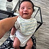 baby, smiling, infant, bib, onesie, baby_clothes, baby_hair, mohawk, person, indoor, floor, glass_table, hand, blanket, heart_pattern, happy, cute, child, seated, home