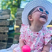 Elise a rejoint le concours — aidez-le/la à gagner de superbes lots ! baby, toddler, child, smiling, laughing, pink_ball, floral_dress, sunhat, sunglasses, outdoor, bench, wood, greenery, daylight, happy, cute, playful, summer, sitting, blanket