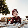 baby, smiling, maroon_clothing, beanie, blanket, outdoor, holiday, christmas_tree, gifts, festive, crawling, child, happy, toy, decorations, nature, sunlight, portrait, cute, seasonal