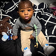 Zavon is registered to the contest to win money with this photo: toddler, child, infant, baby, sitting, indoors, clothing, sweater, pants, sneakers, pacifier, blanket, pattern, expression, face, curious, human, person, toy