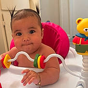 Paloma is registered to the contest to win money with this photo: baby, bracelet, bright_colors, child, chubby_cheeks, closeup, curious_expression, cute, face, hand, indoor, infant, person, pigtails, pink_seat, plastic_toy, play_tray, portrait, toy_rings, yellow_teddy