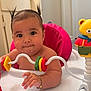 baby, bracelet, bright_colors, child, chubby_cheeks, closeup, curious_expression, cute, face, hand, indoor, infant, person, pigtails, pink_seat, plastic_toy, play_tray, portrait, toy_rings, yellow_teddy