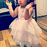 child, girl, dress, pink, flower, headband, clapping, shoes, wooden_floor, blanket, chair, door, indoor, person, toddler, curly_hair, cute, standing, home, floor