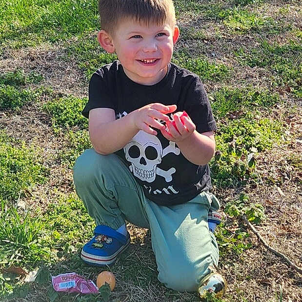 Jaxon joined the competition — help win amazing prizes! child, boy, smiling, outdoor, grass, crouching, skull_tshirt, toy, plastic_eggs, candy, sunlight, greenery, playful, happy, casual_clothing, blue_shoes, nature, daylight, young, fun