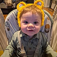 Barney is registered to the contest to win money with this photo: toddler, child, baby, high_chair, yellow_ears, fuzzy_headband, face, indoor, dog, pet, couch, living_room, curious, straps, clothing, person, smile, home, cute, seated