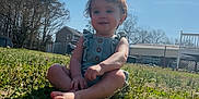 Rhyea is registered to the contest to win money with this photo: child, toddler, smile, barefoot, grass, outdoor, sunlight, romper, yard, house, fence, trees, blue_sky, portrait, sitting, play, nature, weekend, young_child, happy