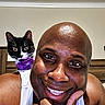 Pepper is registered to the contest to win money with this photo: background_wall, bald, black_and_white_cat, cat, close_up, eyes, flower_collar, hand, happy, indoor, man, portrait, purple_flower, ring, shoulder, skin, smiling, tank_top, whiskers, wooden_headboard