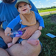 Rhett is registered to the contest to win money with this photo: adult, baby, beard, bib, blue_shirt, casual_clothing, child, clouds, grass, green_cap, happy, holding, legs, nature, outdoor, phone, sky, smile, sunny, water_bottle