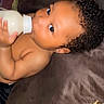 baby, bottle, milk, curly_hair, cushion, lying_down, infant, skin, hand, face, child, indoors, soft_texture, closeup, feeding, cute, expression, relaxation, comfort, toddler