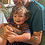 adult, baby, child, tattoos, curly_hair, smiling, indoor, seated, person, holding, clothing, striped_shirt, blue_shorts, furniture, home, window, natural_light, happy, family, care