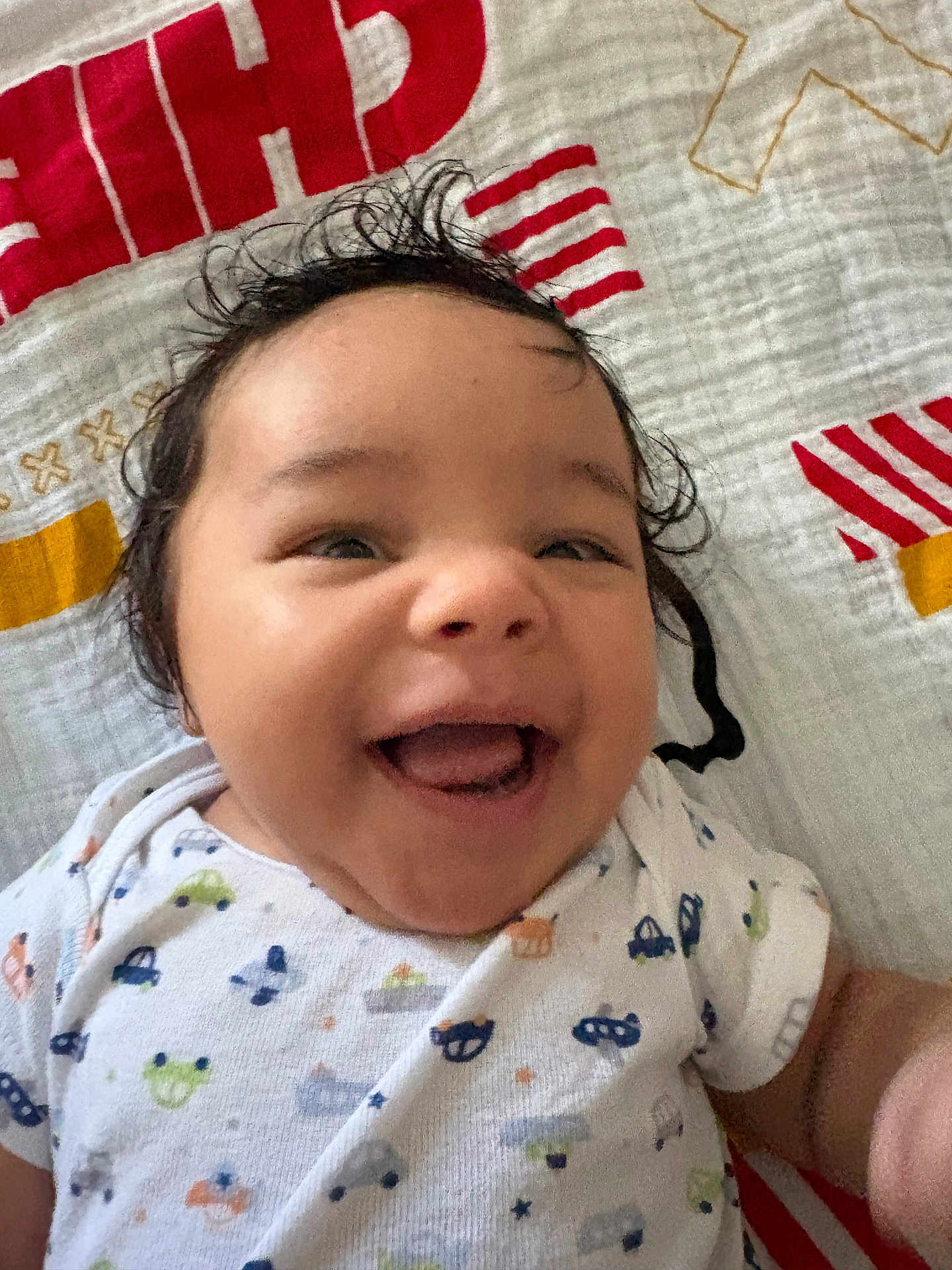 Zechariah joined the competition — help win amazing prizes! baby, smiling, happy, infant, face, portrait, clothing, onesie, patterned_clothing, lying_down, blanket, curly_hair, cute, child, person, indoor, closeup, skin, joy, expression