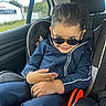 child, car_seat, sunglasses, blue_tracksuit, car_interior, seat_belt, smile, hair, hand, window, trees, daylight, road_sign, casual_clothing, cute, portrait, young_child, playful, confident, car