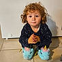 appliance_background, blue_unicorn_slippers, brown_eyes, candid, child, curly_hair, cute, hands_together, home, indoor, looking_up, navy_robe, neutral_expression, plush, portrait, shadow, sitting, slippers, tiled_floor, toddler