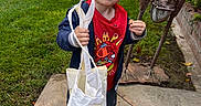 Caleb is registered to the contest to win money with this photo: child, toddler, boy, portrait, outdoor, garden, lawn, smiling, snack, food, tote_bag, sling_bag, red_shirt, jacket, sandals, garden_sculpture, brick_wall, patio, greenery, daylight