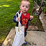 child, toddler, boy, portrait, outdoor, garden, lawn, smiling, snack, food, tote_bag, sling_bag, red_shirt, jacket, sandals, garden_sculpture, brick_wall, patio, greenery, daylight
