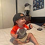 toddler, child, bed, cap, orange_shirt, shorts, smile, indoor, lamp, wall, photocollage, furniture, room, person, barefoot, sitting, paper, headwear, young_child, casual