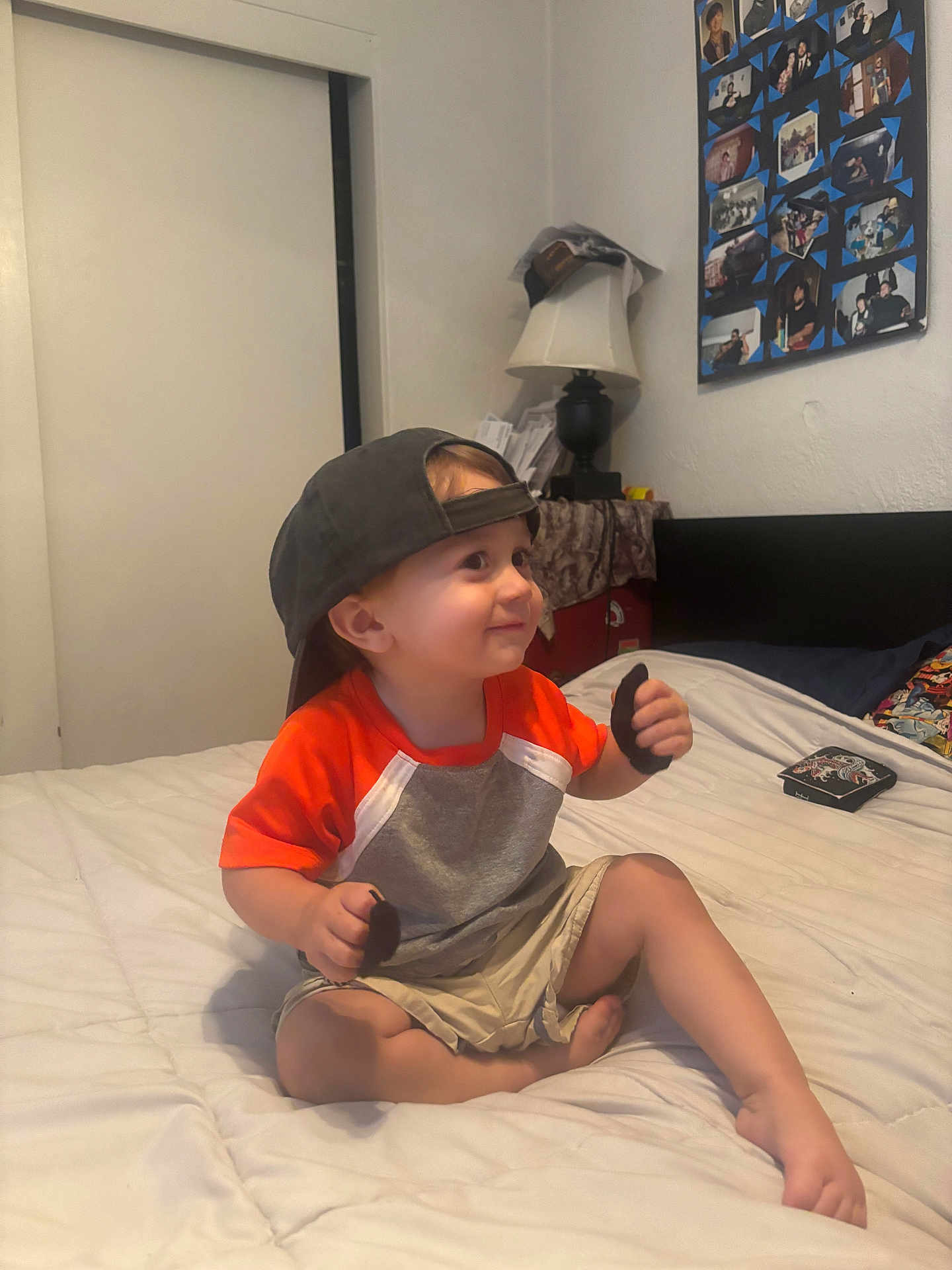 Caleb is registered to the contest to win money with this photo: toddler, child, bed, cap, orange_shirt, shorts, smile, indoor, lamp, wall, photocollage, furniture, room, person, barefoot, sitting, paper, headwear, young_child, casual