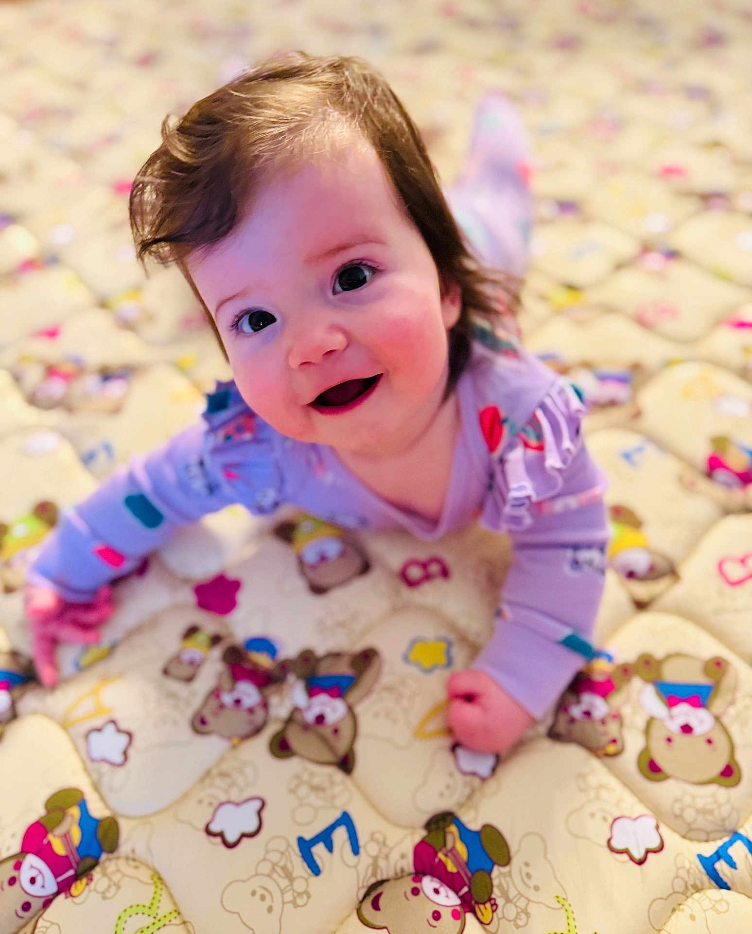 Adele is registered to the contest to win money with this photo: baby, child, smiling, crawling, purple_clothing, quilt, teddy_bear_pattern, indoor, cute, face, happy, infant, playful, young_child, person, soft_lighting, closeup, head, cheerful, colorful