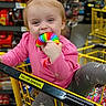 aisle, baby, chewing, child, clothing, cute, grey_pants, grocery_store, happy, indoor, person, pink_shirt, playing, rainbow, retail, shopping_cart, store, toddler, toy, yellow_cart