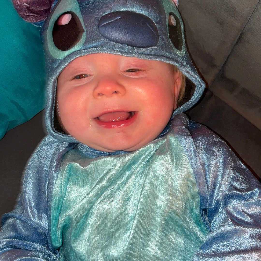 Maximus is registered to the contest to win money with this photo: baby, infant, child, stitch_costume, costume, blue, smiling, happy, portrait, face, eyes, mouth, chubby_cheeks, sitting, indoors, couch, blanket, toy, headwear, photography