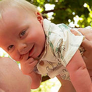 Maximus is registered to the contest to win money with this photo: baby, child, infant, smiling, happy, blond_hair, blue_eyes, portrait, outdoors, sunlight, bokeh, greenery, adult, parent, being_held, hands, finger_in_mouth, white_clothing, family, close_up