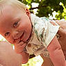baby, child, infant, smiling, happy, blond_hair, blue_eyes, portrait, outdoors, sunlight, bokeh, greenery, adult, parent, being_held, hands, finger_in_mouth, white_clothing, family, close_up