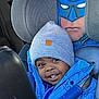child, toddler, car_seat, blue_jacket, gray_beanie, batman_cover, car_interior, smiling, seatbelt, apple, happy, young_child, portrait, winter_clothing, cute, kid, car_window, daylight, seat_headrest, travel