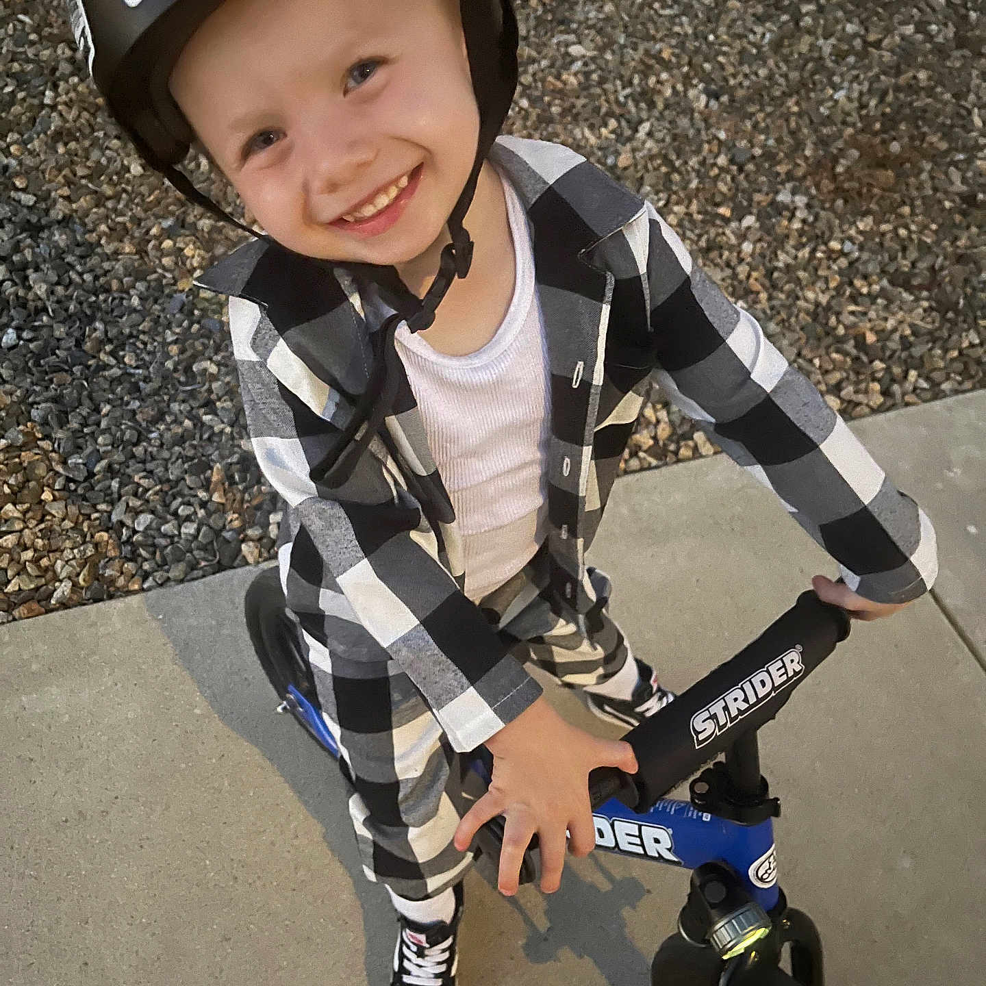 Jason Jr is registered to the contest to win money with this photo: balance_bike, bike, black_and_white_clothing, casual_wear, checkered_pattern, child, daylight, gravel, happy, headgear, helmet, outdoor, person, play, portrait, sidewalk, smiling, sneakers, strider, young_child