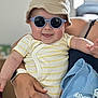 baby, child, sunglasses, hat, onesie, striped_clothing, adult_hand, ring, smartwatch, blue_cloth, tongue_out, smiling, indoor, person, holding, cute, happy, face, headwear, casual