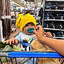 toddler, child, shopping_cart, yellow_hat, pacifier, plush_toy, teddy_bear, hand, clothing, store_aisle, belts, shelves, jeans, retail, shopping, baby, cute, indoor, person, casual