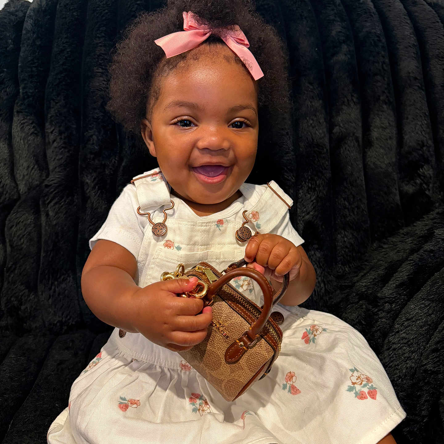 Kadale is registered to the contest to win money with this photo: baby, child, infant, portrait, smiling, happy, bow, pink_bow, curly_hair, black_blanket, dress, floral_dress, overalls, handbag, purse, fashion, sitting, indoors, closeup, photography
