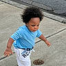 child, toddler, walking, outdoor, sidewalk, street, portrait, blue_shirt, white_shorts, curly_hair, afro_hair, concentrated, determined, standing, movement, pavement, urban, daylight, clothing, manhole_cover