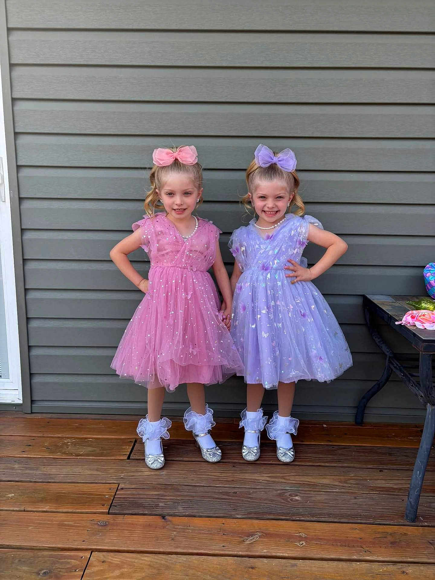 Avalinaandandilyn is registered to the contest to win money with this photo: child, children, girl, twins, dress, pink_dress, purple_dress, bow, hair_bow, smile, pose, shoes, socks, wooden_deck, outdoor, wall, happy, holding_hands, pearls, fashion