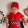 automotive, baby, baby_photography, celebration, checkered_pattern, child, flat_lay, formula_one, headwear, indoor, infant, portrait, race_car, racing_costume, racing_theme, red_outfit, route_66, smiling, studio_photo, toy_car