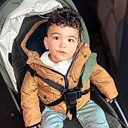 Adam participe au concours pour gagner de l'argent avec cette photo : child, toddler, stroller, jacket, harness, buckle, curly_hair, eyes, face, portrait, sitting, cozy, hood, denim_pants, shoe, hand, zipper, curious_expression, blurred_background, seat