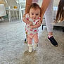 baby, child, person, indoor, carpet, clothing, shoes, hands, legs, furniture, table, chair, window, toy, pacifier, adult, walking, support, home, cute