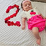 baby, infant, pink_clothing, white_blanket, rose_petals, number_two, headband, cherry_patch, cute, smiling, lying_down, celebration, milestone, soft_texture, child, person, indoors, foot, hand, face