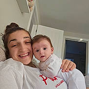 Olivia is registered to the contest to win money with this photo: woman, baby, smiling, indoor, couch, white_clothing, face, hand, hair, door, shelf, toy, teddy_bear, expression, person, portrait, happy, skin, living_room, casual