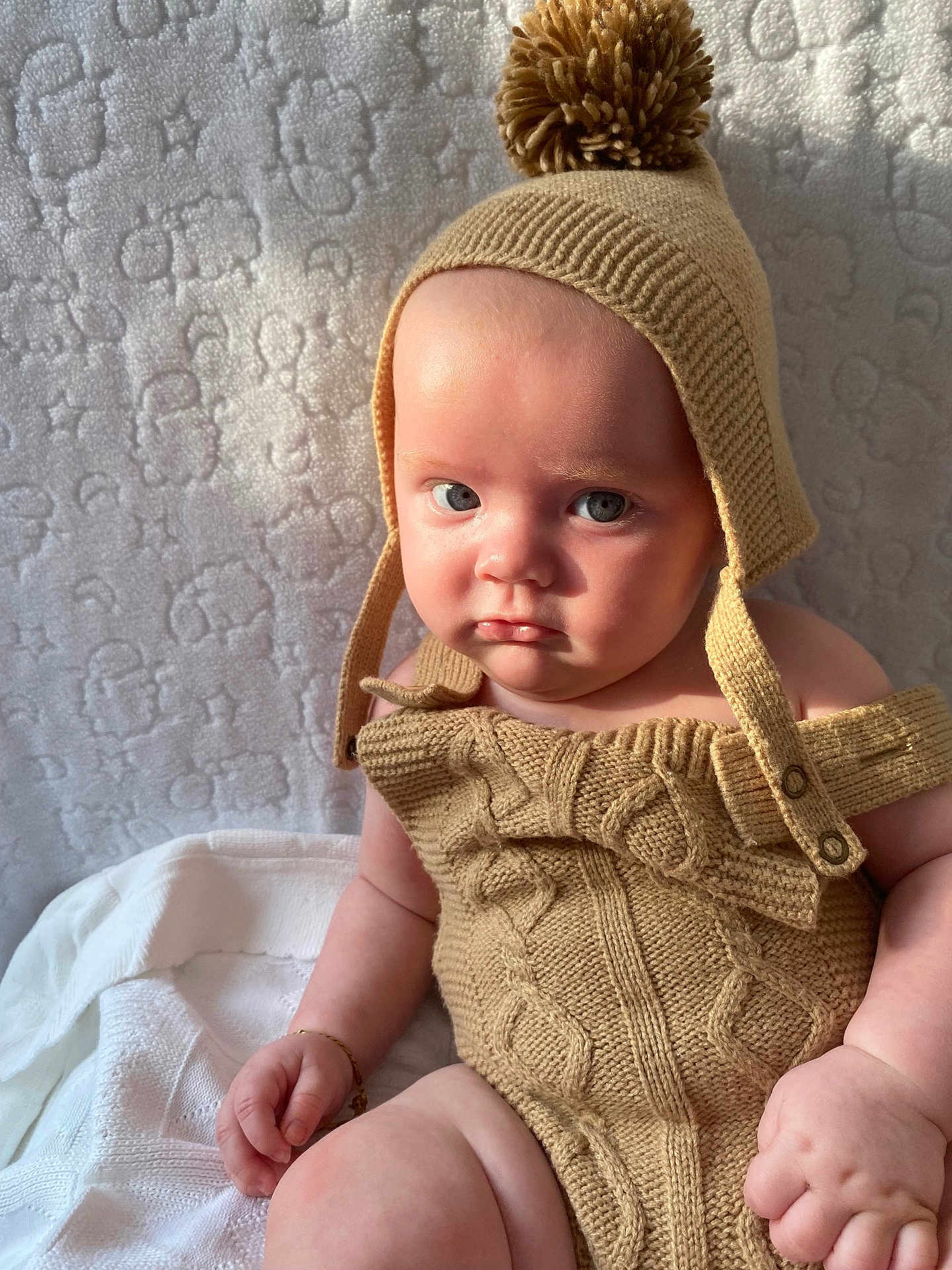 Koa is registered to the contest to win money with this photo: baby, child, knitwear, hat, pom_pom, beige, texture, blanket, portrait, indoor, cute, infant, soft_light, expression, hands, skin, sitting, warm_clothing, closeup, person